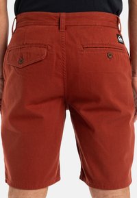 Rust-colored shorts made of cotton, featuring two back pockets with button closures and a branded patch on the waistband.
