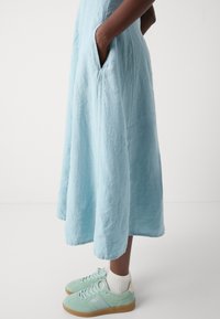 Personne portant une robe midi bleu clair avec des poches, associée à des chaussettes blanches et des baskets vert pastel avec des semelles en gomme.