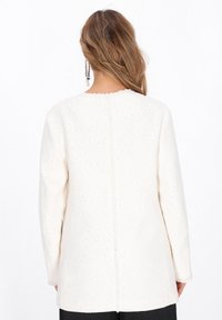 Jachetă albă texturată cu guler rotund, mâneci lungi și embellishments subtile. Spatele este neted, cu o cusătură centrală.