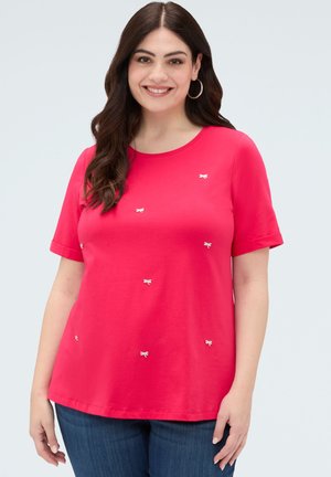 Uśmiechnięta kobieta z długimi ciemnymi włosami nosi jasnoróżową bluzkę z krótkim rękawem, ozdobioną małymi haftowanymi kokardkami, oraz niebieskie dżinsy na jednolitym tle.