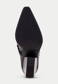 Zapato de tacón alto de cuero negro con punta afilada, suela texturizada, acentos plateados en el tacón y un patrón geométrico distintivo en la parte inferior.