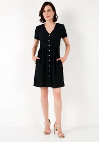 Vestito nero a metà coscia con maniche corte, scollo a V, frontale con bottoni e tasche laterali. Tessuto morbido con una vestibilità rilassata. Bottoni metallici decorativi.