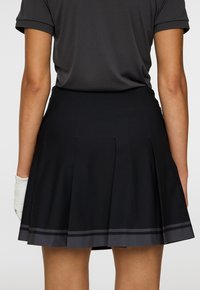 Persoon in een zwarte geplooide rok met dubbele donkergrijze strepen dicht bij de zoom en een zwarte golfhandschoen aan de linkerkant, gezien van achteren.