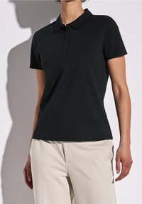 Svart polo shirt med kort ärm, knapprad, mjuk bomullstyg och avslappnad passform, i kombination med ljusbeiga byxor.