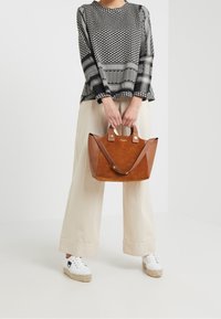Borsa a spalla in pelle marrone con texture scamosciata, accenti neri in contrasto e doppio manico, tenuta da una persona che indossa un top con motivo in bianco e nero.