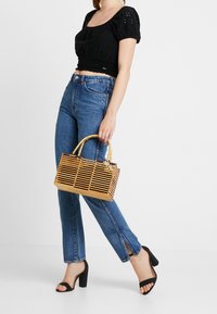 Top court noir à manches ballon, jean bleu taille haute avec ourlet fendu, et petit sac tissé beige avec poignée en bambou.