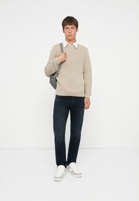 Beige geribbeld sweater met een ronde hals, grijze schoudertas, donkerblauwe jeans en witte sneakers met beige accenten.