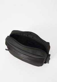 Borsa nera con zip e texture morbida, dotata di una tracolla con logo in evidenza e un ampio interno foderato in tessuto.