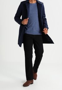 Marinefärgad trenchcoat över en blåtexturerad t-shirt, kombinerad med svarta byxor och bruna stövlar. Trenchcoaten har knapprar och en krage.