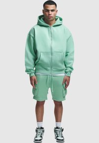 Mentazöld zip-es kapucnis pulóver elülső zsebekkel és hozzáillő cargo sorttal. Fehér zoknikkal és fekete-fehér sneakerrel viselve. Egyszerű, laza szabású.