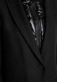 Manteau en laine noire avec de larges revers superposé sur une doudoune noire brillante, zippée à mi-hauteur au niveau de la poitrine.