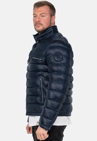 Giacca puffer in pelle blu navy con colletto alto, tasca sul petto con zip e design trapuntato testurizzato. Presenta il logo sulla manica.