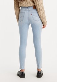 Jeans skinny de color azul claro, hechos de mezclilla, con bolsillos traseros y una etiqueta de cuero marrón en la cinturilla. Llevados con zapatos negros.