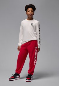 Pika varrukatega valge särk logoga, punased dressipüksid tekstiga „JORDAN” ning mustade ja punaste spordijalatsitega. Sile kangas, lõdvestunud istuvus, sportlik stiil.