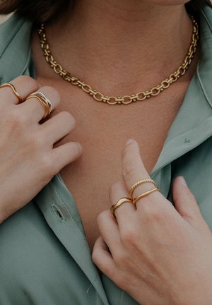 Zelta ķēdes kaklarota ar ovālas formas saitēm. Dažādi zelta gredzeni ar vijumiem un gludu dizainu. Redzama gaiši zaļa blūze.