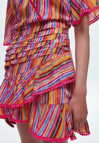 Top multicolore a righe con rouches sovrapposte e rifiniture in pompon rosa. Il materiale appare leggero e con una consistenza particolare. Palette di colori brillante e vivace.