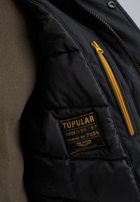 Črna puhovka z rumenim zadrgo žepom, ki ima tkaninsko nalepko z oznako "TUPULAR" in izrazito teksturo. Vidna mehka podloga.