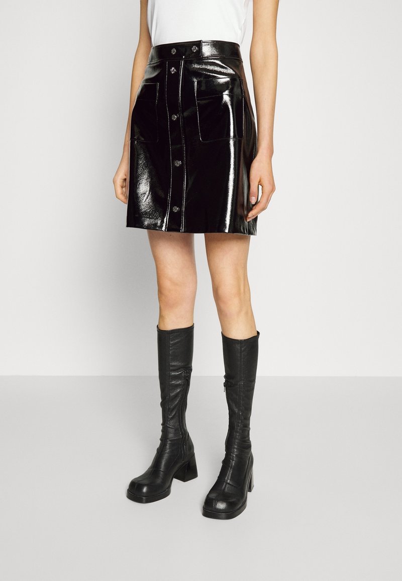 Femme portant une mini-jupe noire brillante boutonnée avec de grandes poches et des bottes noires à talons montant jusqu’aux genoux, sur un fond uni.