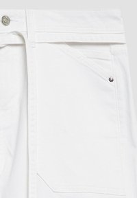 Gros plan sur le côté avant gauche d'un jean en denim blanc montrant les détails de la poche, de la passants de ceinture, du bouton et des rivets.