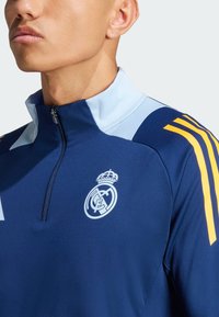 Tengerészkék negyedcipzáros edzőfelső világoskék gallérral, fehér Real Madrid logóval és három sárga csíkkal a jobb vállon.