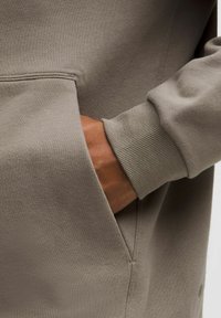 Hellgrauer Sweatshirt-Stoff mit sichtbarem Fronttasche und gerippten Bündchen. Eine Nahaufnahme zeigt die glatte Textur und präzise Nähdetails.
