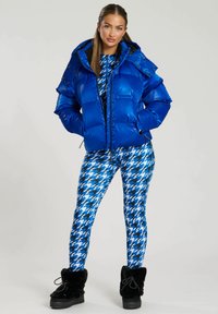 Blå puffjacka med blanka ytor, knäppning fram och huva, matchad med åtsittande leggings i blått och svart houndstooth-mönster samt svarta stövlar med pälsfoder.