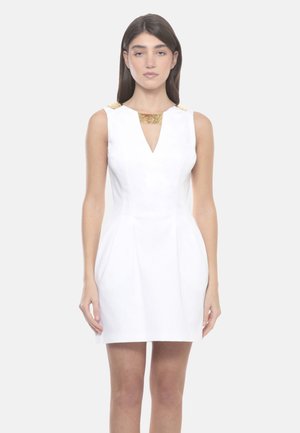 Femme debout portant une robe blanche sans manches avec des accents dorés sur les épaules et l'encolure, sur un fond clair uni.