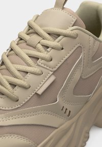 Zapatilla deportiva beige gruesa con paneles de malla y cuero, detalles cosidos, suela gruesa y cordones beige apretados.