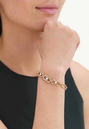 Gouden kettingarmband met ovale schakels, elk met een witte parel, gedragen om de pols bij een zwarte mouwloze top.