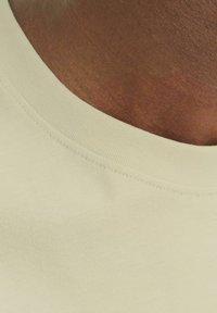 Hellbeige T-Shirt mit rundem Halsausschnitt aus Rippstrick und glatter Oberfläche, die den Hautton unter dem Kragen zeigt.