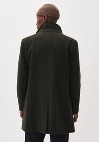 Manteau en laine vert foncé avec un col montant et une silhouette épurée. Présente une fente centrale au dos et des manches droites.