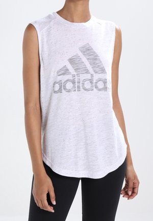 Vrouw draagt een wit mouwloos Adidas topje en zwarte leggings, staand tegen een effen achtergrond.