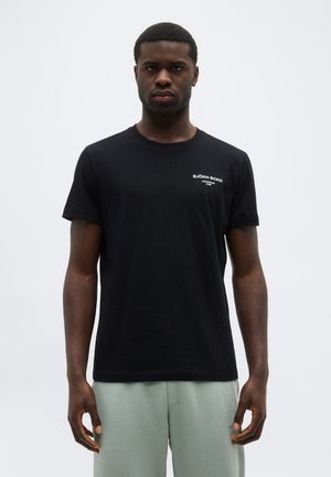 Férfi fekete Björn Borg pólóban és világosszürke nadrágban, egy egyszínű világosszürke háttér előtt áll, előre nézve.