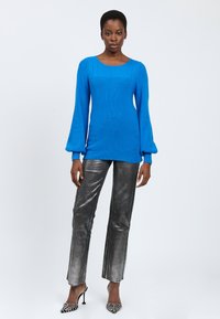 Frau steht in einem leuchtend blauen Strickpullover, metallisch silbernen Hosen und schwarz-weißen High-Heels vor einem schlichten hellen Hintergrund.
