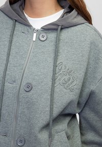 Sudadera con cremallera en gris, con capucha gris a contraste, que presenta dos grandes botones y costuras decorativas en el pecho. Textura de textil suave.
