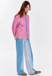 Kobieta ubrana w różowo-czerwoną, w paski kurtkę zapinaną na zamek, szerokie dżinsy w dwóch kolorach oraz mokasyny w panterkę, stojąca z rękami za plecami.