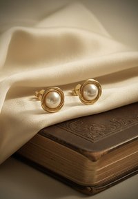 Guldtonade studsörhängen med en rund, vit pärla omgiven av en texturerad cirkulär ram, vilande på krämfärgad tyg över en brun bok.
