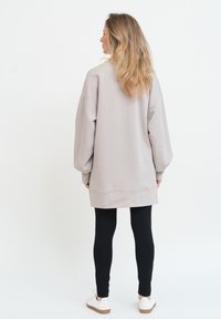 Kvinna med långt blont hår iförd en oversized beige sweatshirt, svarta leggings och vita sneakers som står vänd bort mot en vit bakgrund.