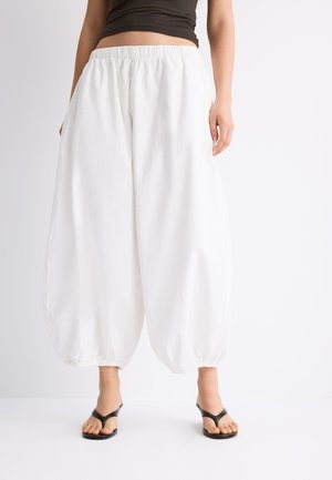 Žena má na sebe voľné biele elastické nohavice na členky, čierny krátky top a čierne sandále s otvorenými prstami, stojí pred jednoduchým bielym pozadím.