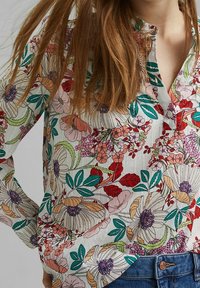 Chemise à manches longues à motifs floraux, mettant en avant des fleurs et des feuilles multicolores sur un fond clair, avec un col boutonné et un tissu texturé.