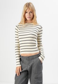 Blondīne sieviete, tērpusies krēmkrāsas garām piedurknēm svīterī ar melnām svītrām un brīvām tumši pelēkām kraukļu biksēm, stāvot uz balta fona.