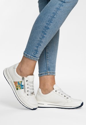 Sneakers bianche con lacci e cerniera laterale, dettaglio colorato e decorato su una scarpa, indossate con jeans azzurri arrotolati.