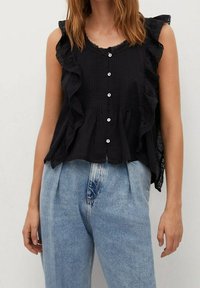 Top nero senza maniche con bordi arricciati, chiusura con bottoni e pizzo. Indossato con jeans a vita alta blu chiaro, che mostrano un contrasto di colori.