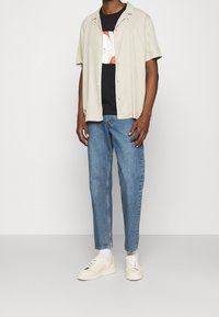 Mand iført beige kortærmet skjorte med knap foran over en sort grafisk T-shirt, blå jeans, hvide sokker og cremede sneakers, stående foran en hvid baggrund.