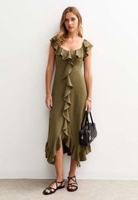 Vestido midi de color verde oliva con detalles de volantes en el escote y a los lados, tejido texturizado, acompañado de un bolso negro y sandalias.