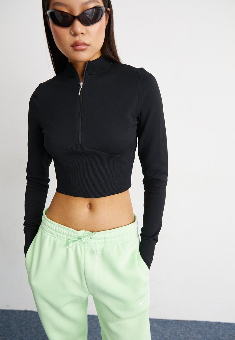 Schwarzes langärmeliges Crop-Top mit einem Reißverschluss vorne, kombiniert mit hellgrünen Sweatpants mit Kordelzug. Glatte Textur und enganliegendes Design.