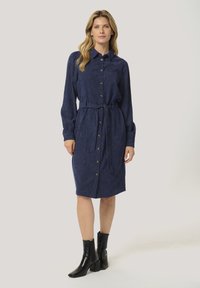 Robe chemise bleu marine à boutons en tissu texturé avec une taille ceinturée, des manches longues et des boutons dorés. Portée avec des bottines noires.