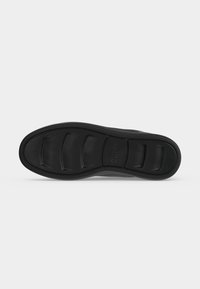 Semelle de sneaker noire avec des motifs rectangulaires en relief et le nom de la marque "Guess" embossé au centre.