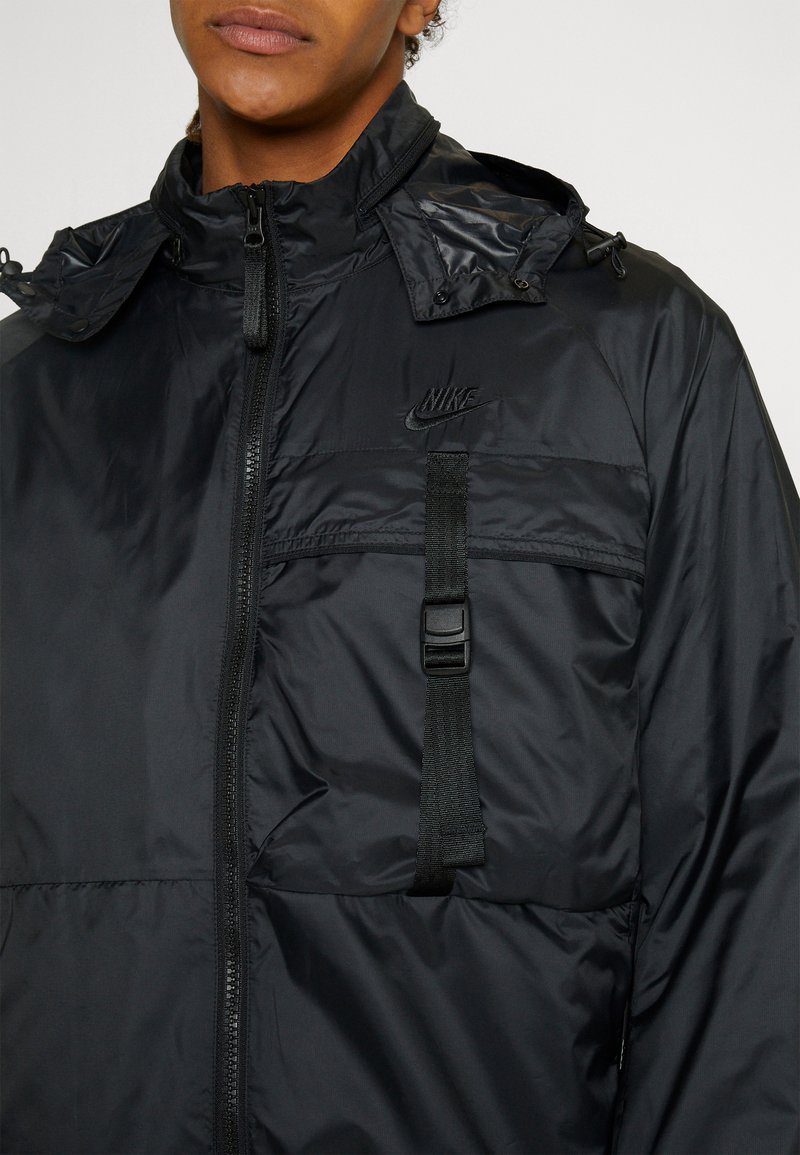 Giacca nera Nike con zip frontale, colletto alto e cappuccio regolabile. Presenta una cintura frontale e un logo. Tessuto liscio e lucido.