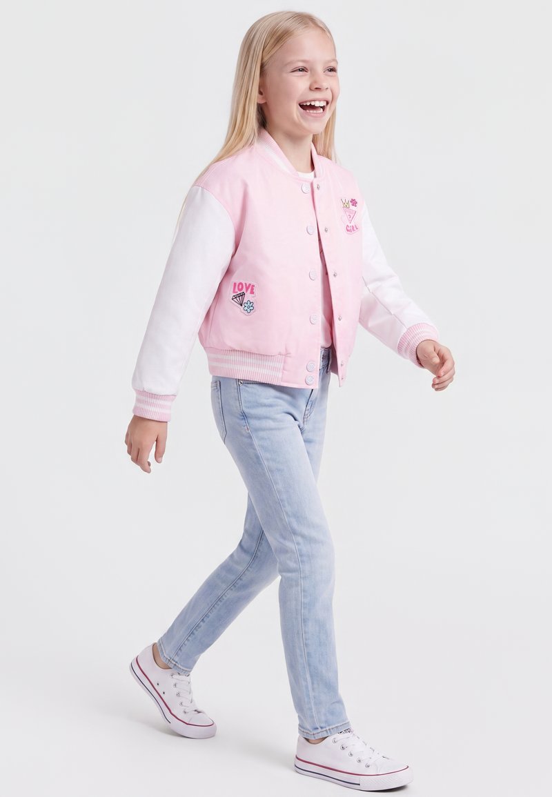 Fille souriante marchant vêtue d'une veste rose et blanche avec des patchs, un jean bleu clair et des baskets blanches sur un fond uni.